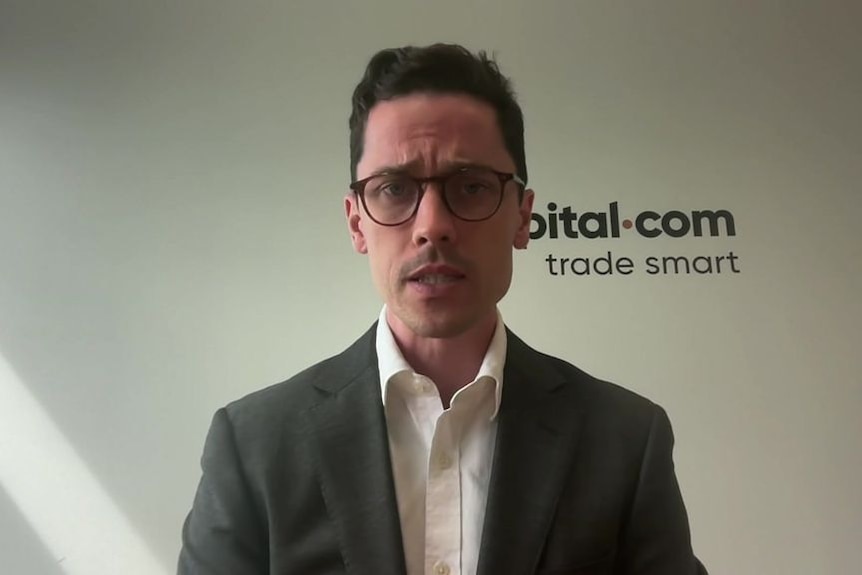 Kyle Rodda wearing a suit and glasses sitting at desk in front of capital.com sign