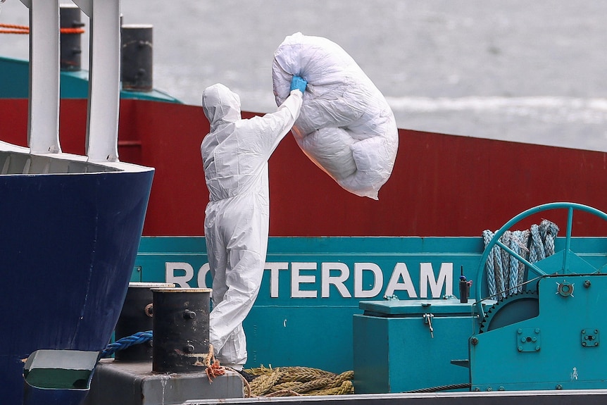 a person in full PPE throws a white bag over the edge of a docked ship