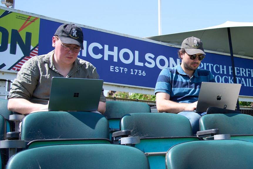 Two young men work on laptops while sitting in the bleachers at a sports ground.