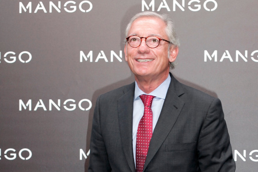 A man with grey hair, glasses, dark suit and red tie, smiles as he stands in front of a wall branded with the Mango logo.