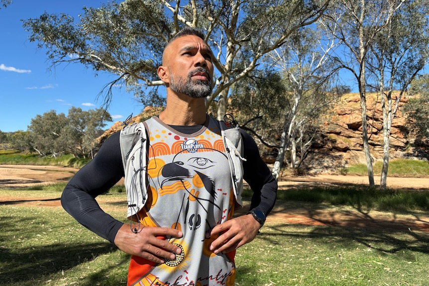 An Aboriginal man wearing black sleeves, a team shirt and a running vest stands with his hands on his hips, looking out.