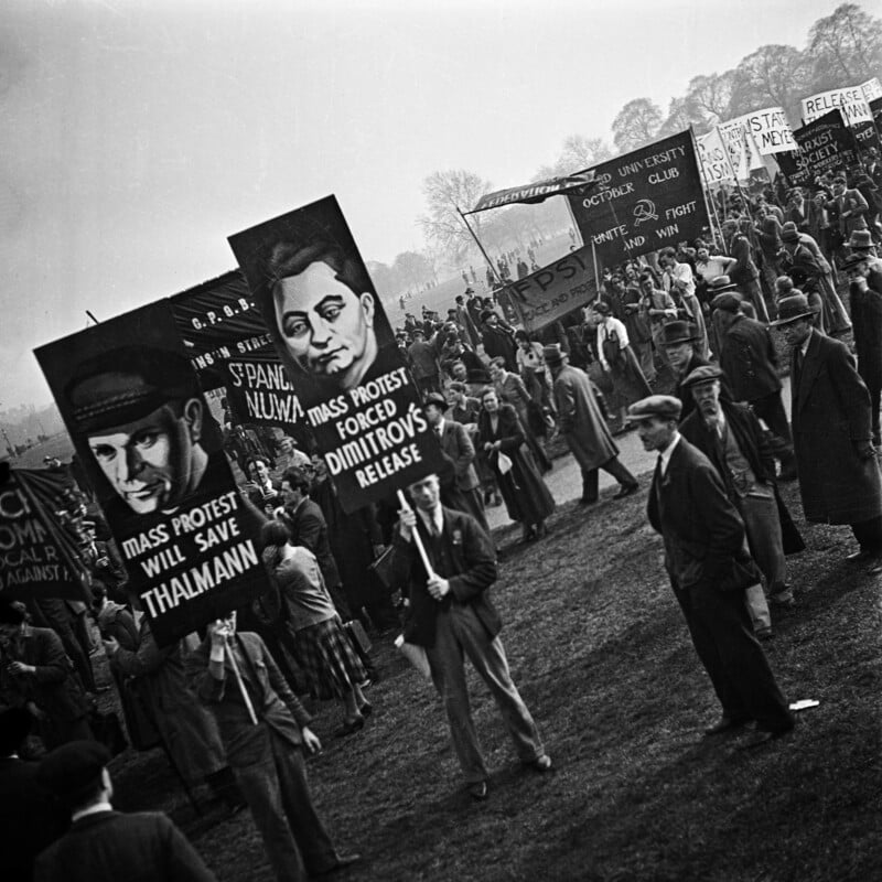 A black-and-white photo of a large outdoor protest with people holding signs featuring illustrated portraits and messages like "Mass protest will save Thalmann" and "Mass protest forced Dimitrov's release.