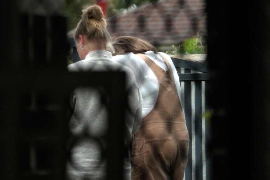 Two women stand their backs to the camera. The picture is taken through a security screen. One woman has her head lowered.