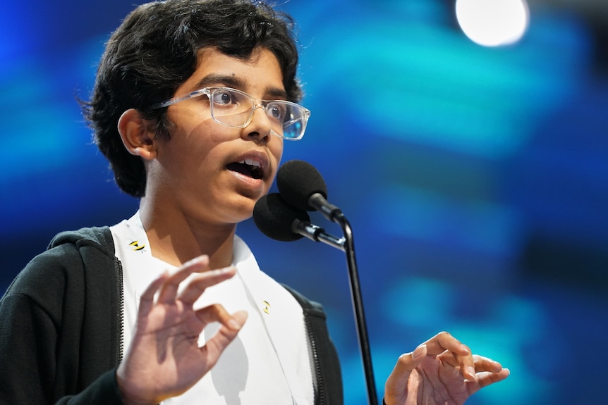 A close up shot of a boy with glasses, talking into a microphone
