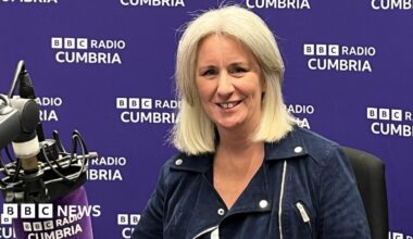 Heather Wainwright sitting in a BBC Radio Cumbria studio, with a branded purple microphone in front of her and a branded purple wall behind her. She has blonde/white hair in a bob and is wearing a blue jacket. She is smiling at the camera.