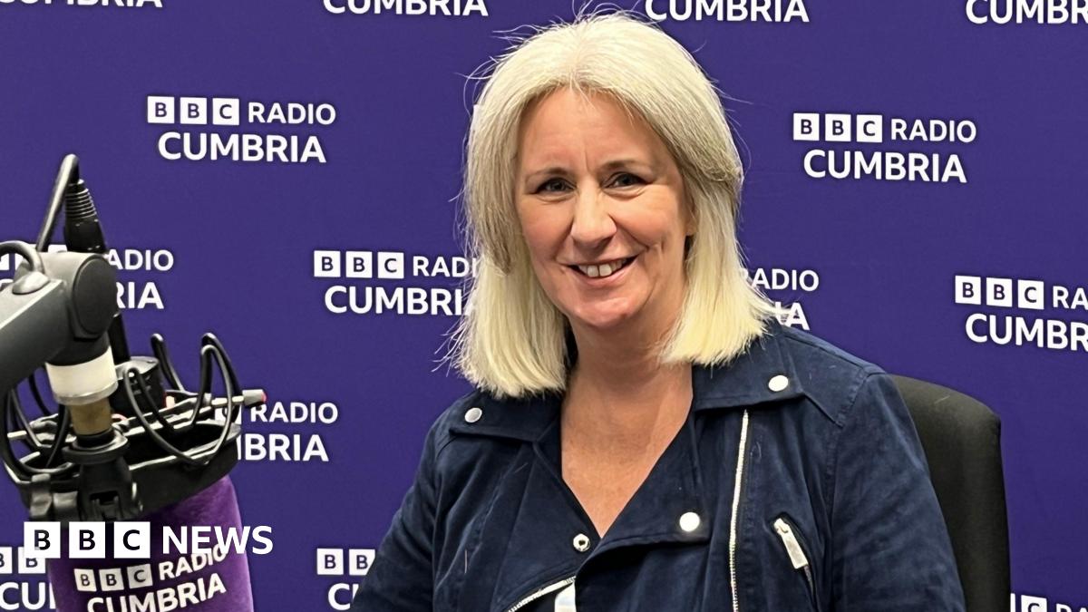 Heather Wainwright sitting in a BBC Radio Cumbria studio, with a branded purple microphone in front of her and a branded purple wall behind her. She has blonde/white hair in a bob and is wearing a blue jacket. She is smiling at the camera.
