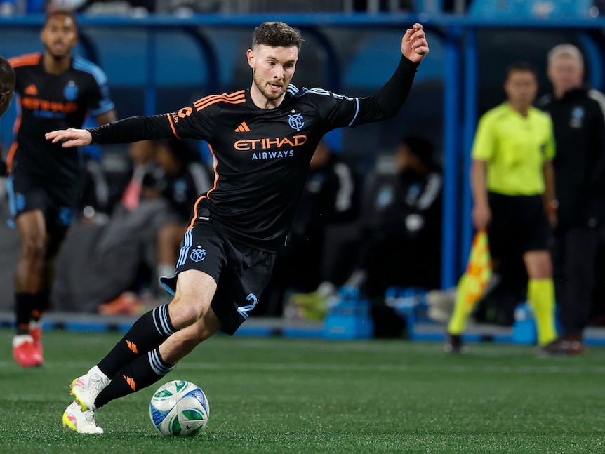 Aiden in Major League Soccer dribbles with the ball while looking upfield, wears black and orange jersey.