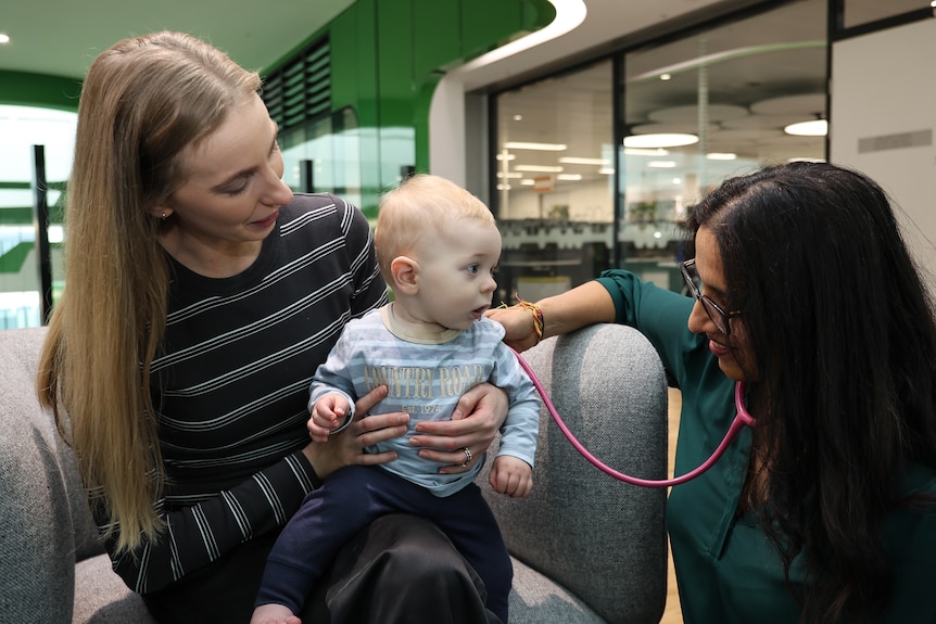 A female doctor holds a stethoscope over a baby being held by his mother