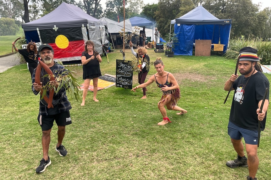 The Aboriginal flag hangs from one of several marquees while a group of people, one holding boomerangs, stand in the foreground.