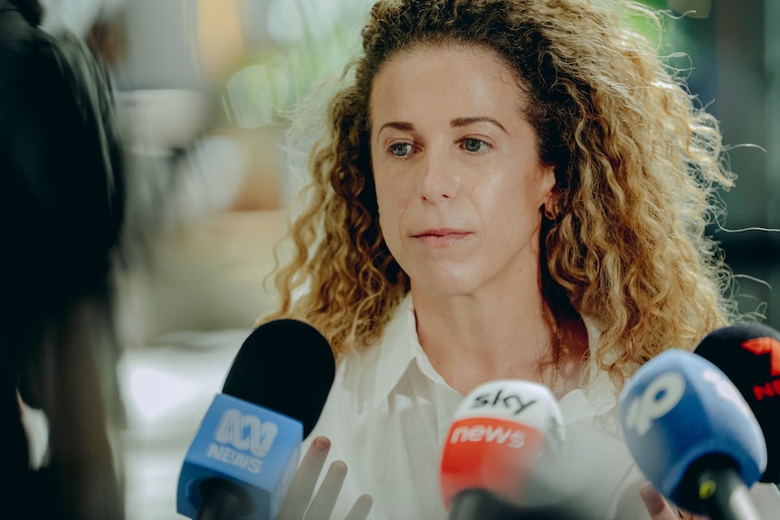 A woman with curly blonde hair wearing a white shirt.