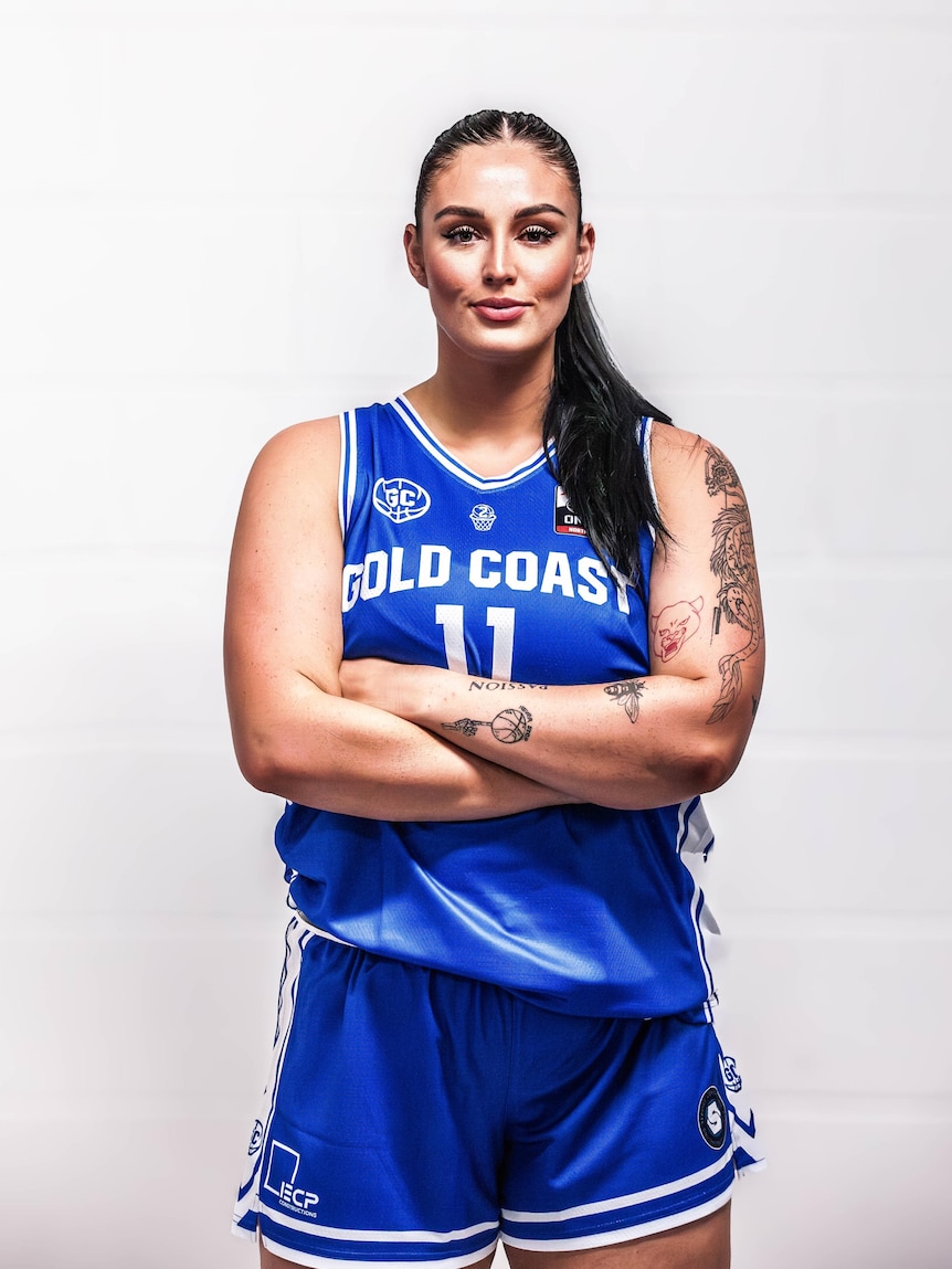 A woman with arms crossed, wearing blue sports jersey and shorts, looking directly at the camera.