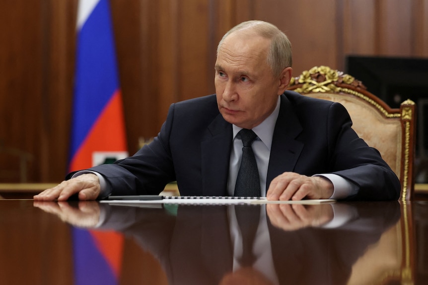 Vladimir Putin sitting at a desk in a meeting room with a Russian flag behind him.