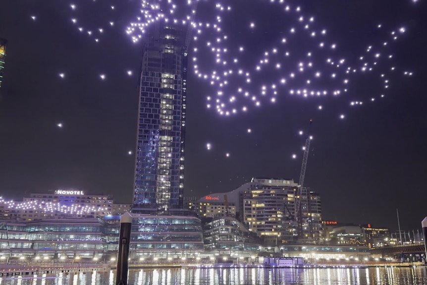 Drone lights falling into the water below, a skyscraper in the background.