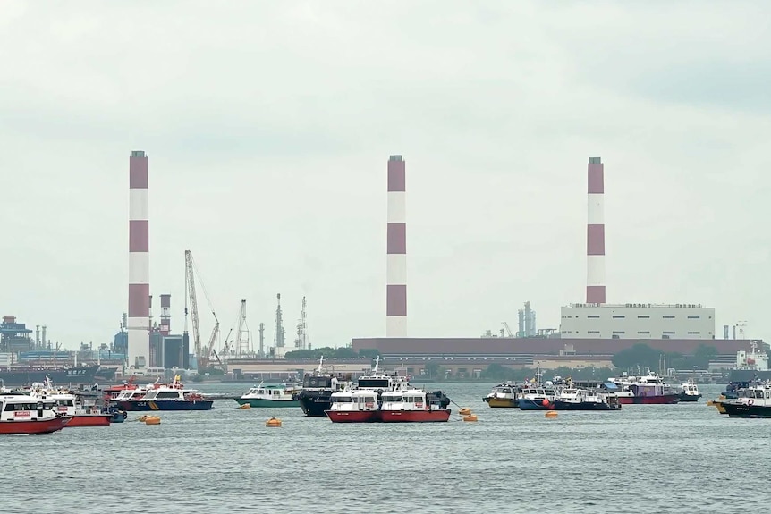 Boats sit in the waters of Singapore, with refinery towers in the background. 