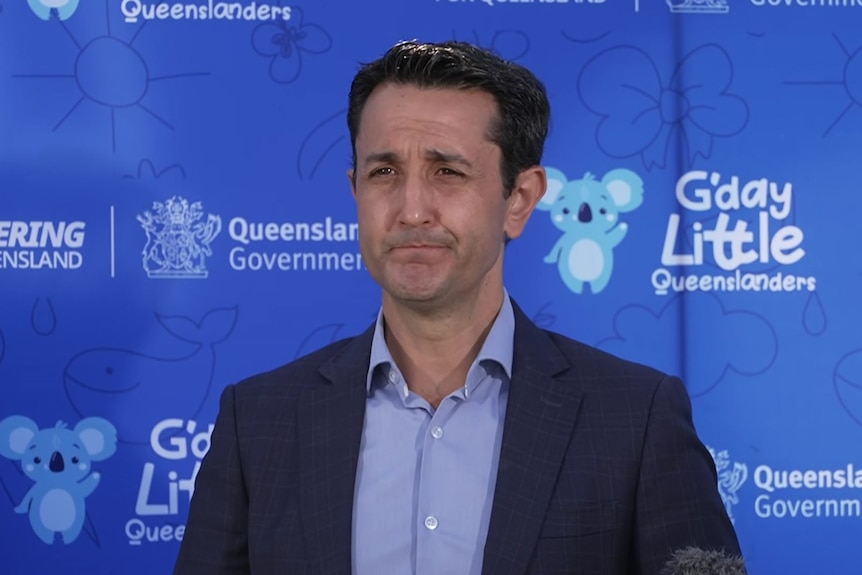 A man in a suit standing on front of blue Queensland government background.