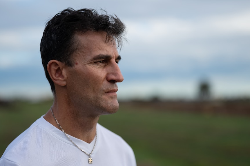 A man in his 40s with cropped dark hair wearing a white t-shirt looks into the distance standing in a green paddock.