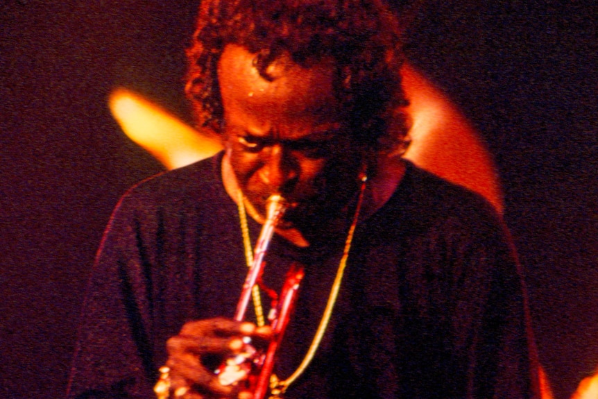 Miles Davis plays his red trumpet on-stage; he's bathed in orange light