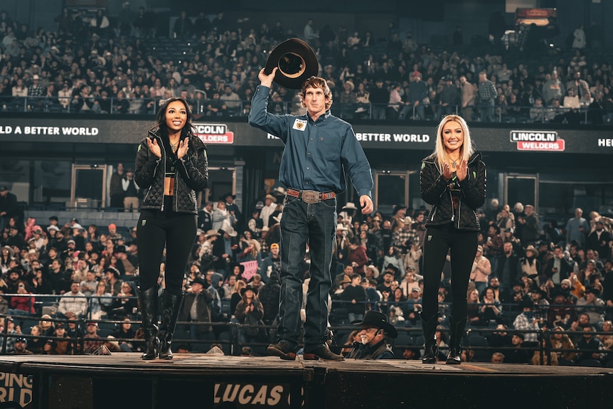 A bull rider standing on a stage with two women as he salutes the crowd with his cowboy hat.