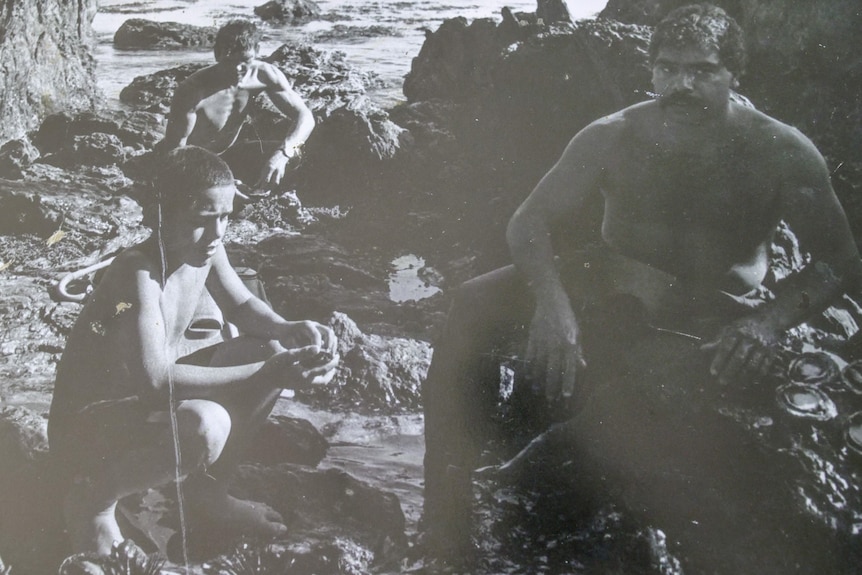 Black and white photo of two men in wetsuits and a young boy with shellfish laid out on the rocks 