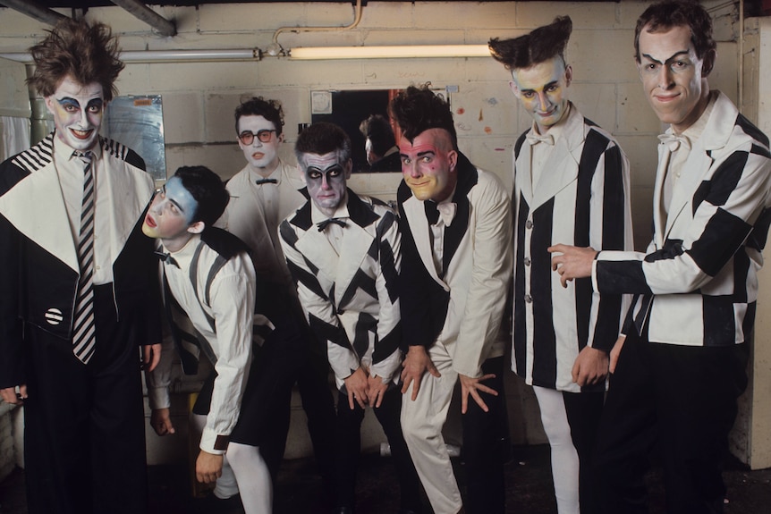 Six members of Split Enz in black‑and‑white outfits with stylized face paint pose backstage near mirrors and pipes.