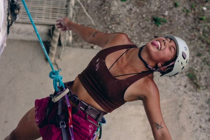 Tanned rock climber woman hanging from a rope and harness, yongue out, eyes closed, leaning back, POV: from above