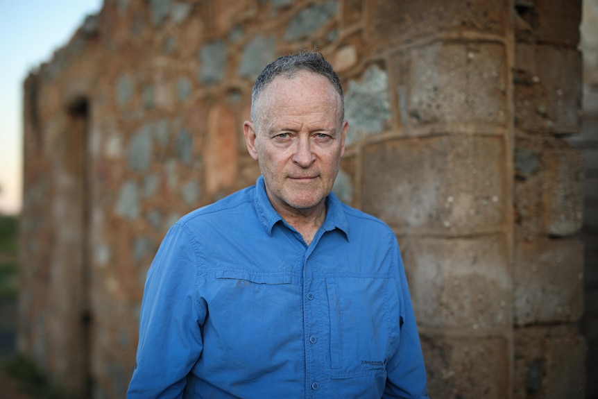A man wearing a blue collared shirt has a stern facial expression. He's standing in front of an old brick building.
