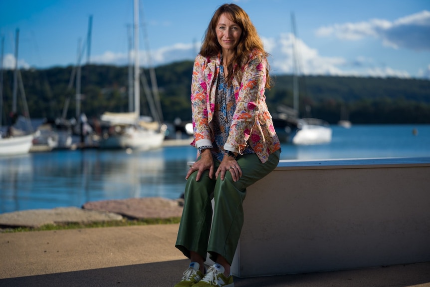 A woman sits on a ledge, behind her is water and boats.