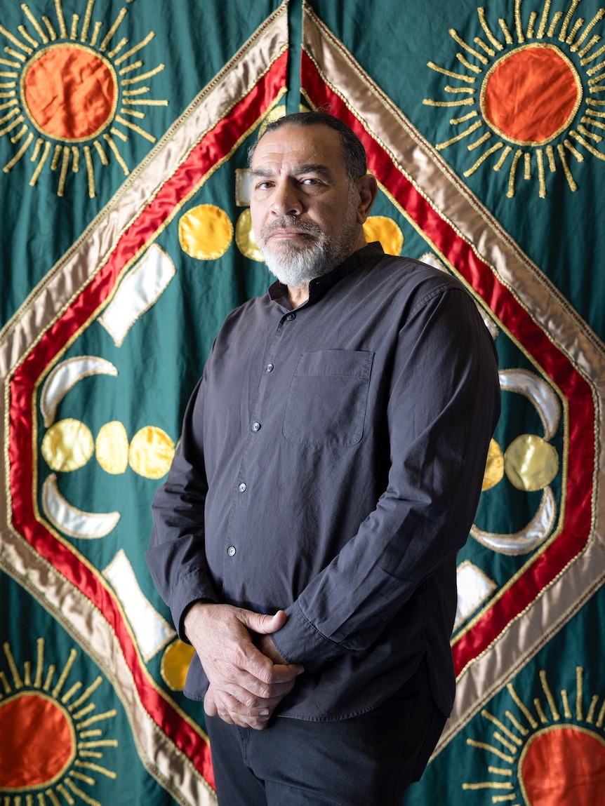 Khaled Sabsabi, a middle-aged Middle Eastern man, hair tied back stands in front of an intimately stitched green tapestry.
