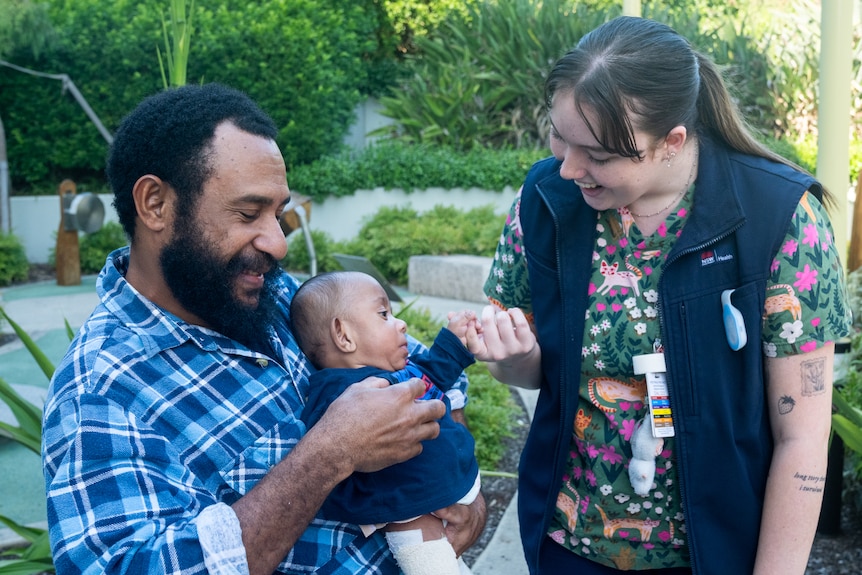 A close up of a man holding a baby, who smiles as he looks up at a woman.