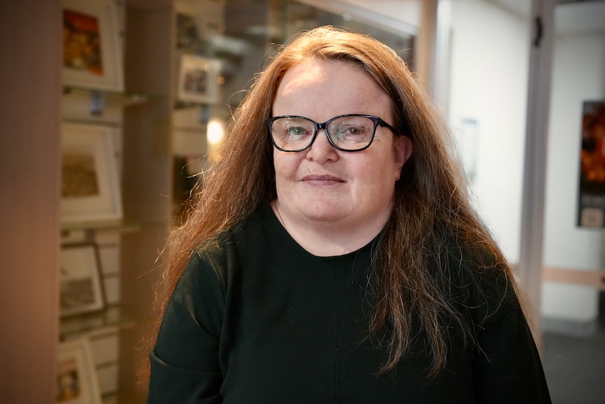 Soneva has glasses and long brown hair and poses for a photo near a display cabinet.