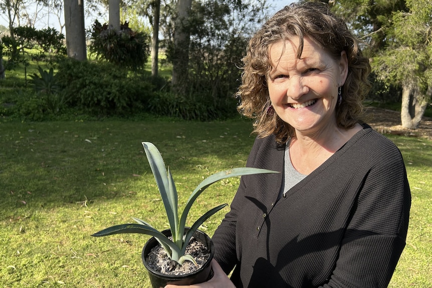 woman in black cardigan smiles to camera holding plant in black pot