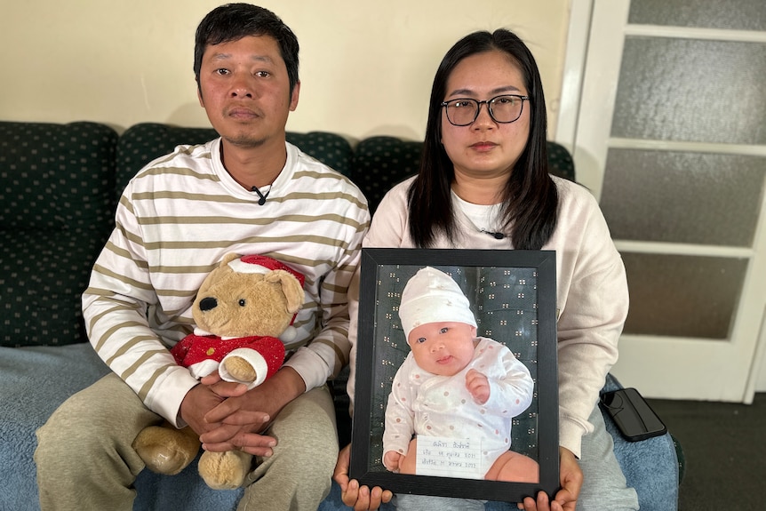 A man in a white and gold striped top holds a teddy while sitting on a couch beside a woman holding a framed photo of a baby.