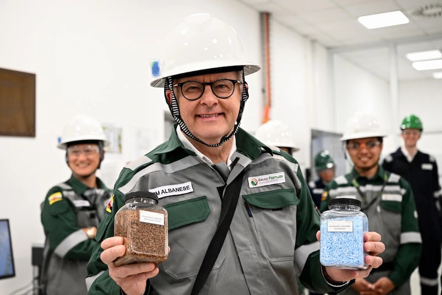 Anthony Albanese in a grey and green jacket and white hard hat, smiling while holding up two jars of blue and brown urea.
