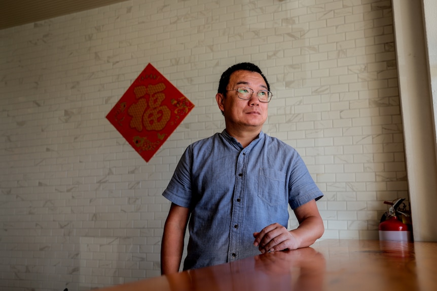 Man wearing glasses looks out shop window with red Chinese sign pinned to white brick wall
