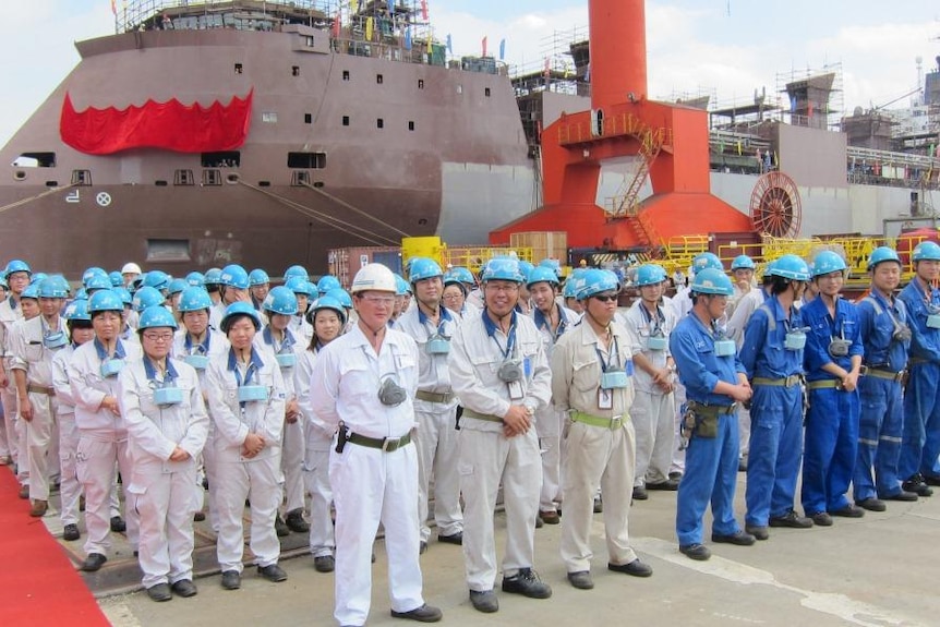 Workers in hard hats and coveralls stand in front of a docked ship.