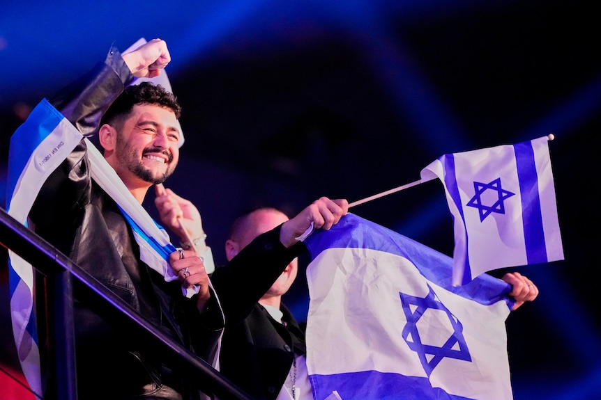 A man smiles and raises a fist while Israeli flags are waved around him