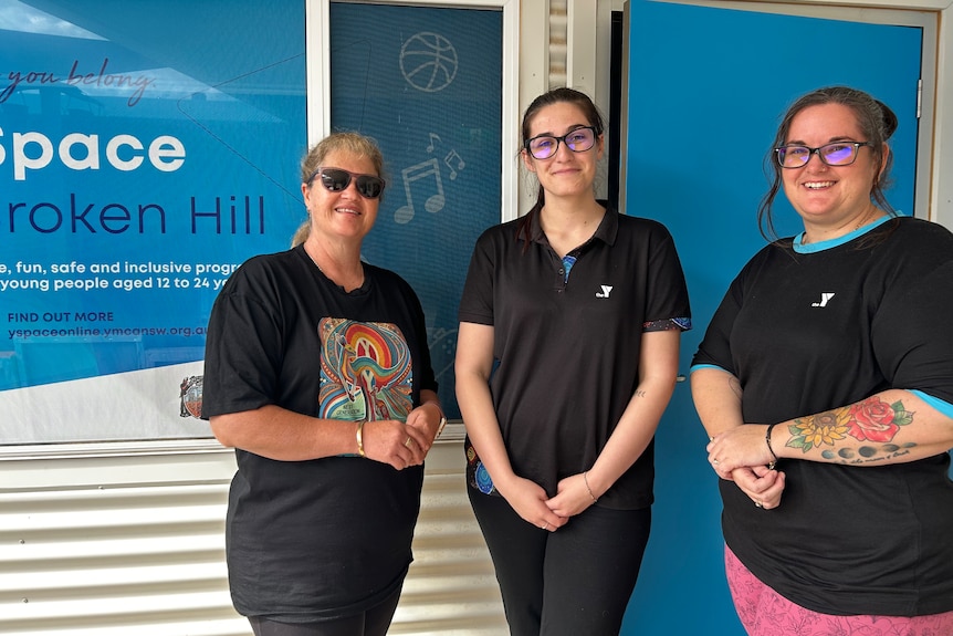 Three women wearing The Y uniforms standing in front of a blue sign and smiling at the camera
