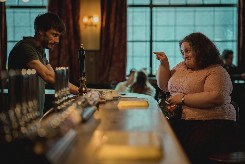 two people are looking at each other, a man is standing at a bar smiling, the woman is sitting, smiling and pointing at him.