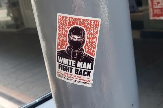 A sticker depicting a hooded character with the text "White man fight back" is stuck to a pole on a street.