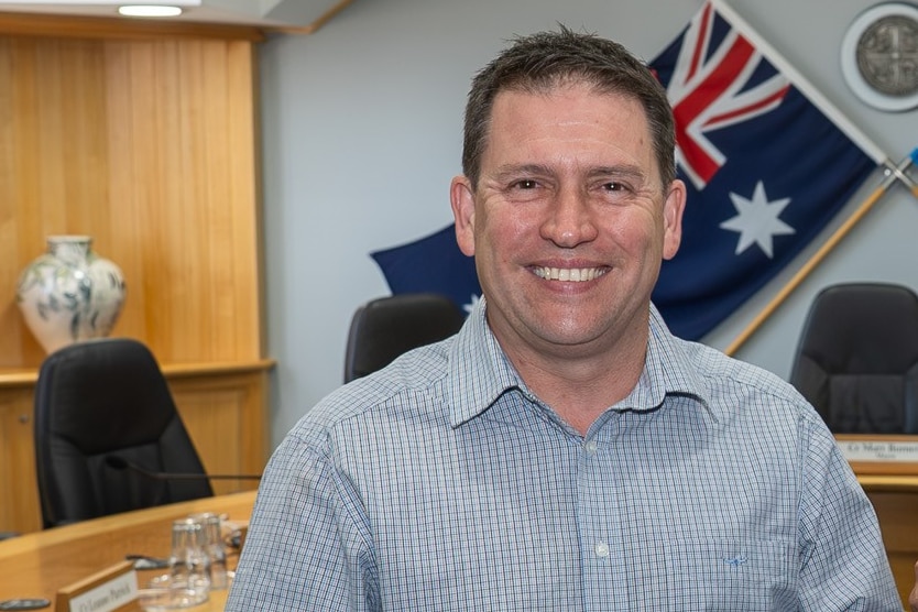 A man with dark hair smiles, he is standing in council chambers with an Australian flag behind him