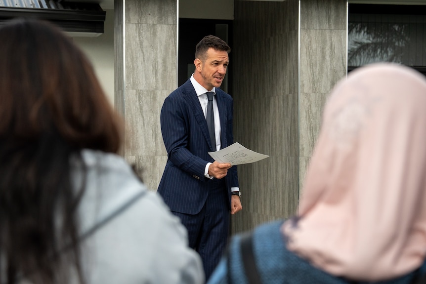A man in a suit holding a piece of paper outside in front of groups of people waiting looking at him.