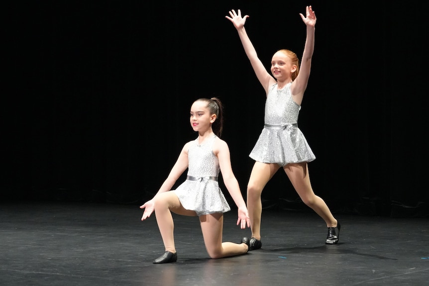 Two little girls in white jazz costumes strike a pose against a black stage. One has her hands up standing behind the other.