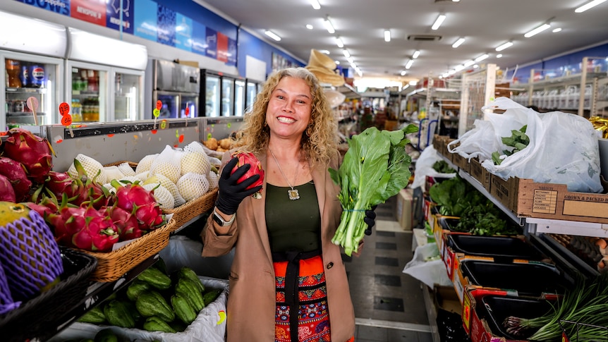 Woman with curly hair smiles holding fruit and vegetable standing inside Asian grocer supermarket