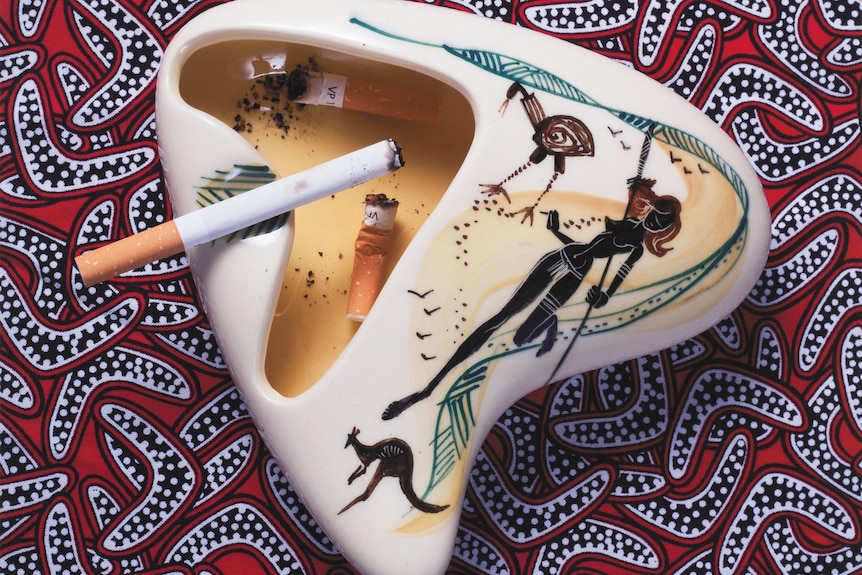 An ash tray with black figure painted onto it and cigarette sitting on it, on material with boomerangs printed on it.