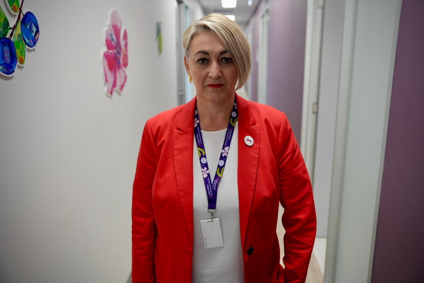 A woman in a red blazer and white shirt and short blonde hair stands in a hallway.