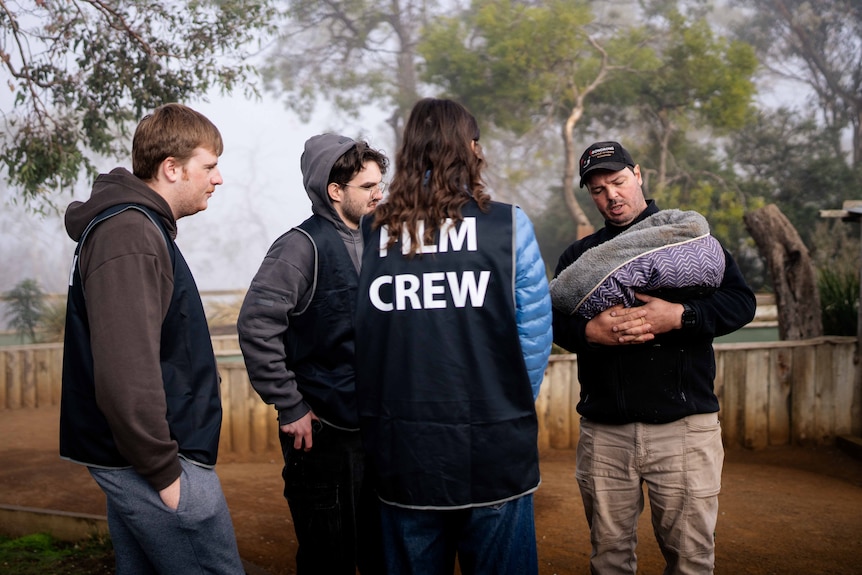 Four people in a huddle at a wildlife sanctuary. Oner wears a vest that says 'film crew' and another holds a wombat.