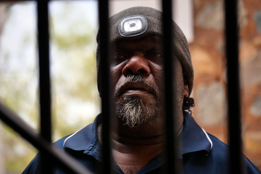 An Aboriginal man with a grey goatee and wearing a beanie stares through prison bars.
