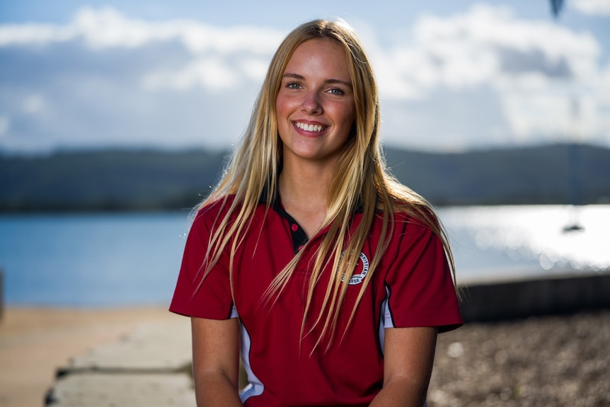 A young woman with blonde hair wearing a red top, sits in the sun and smiles.