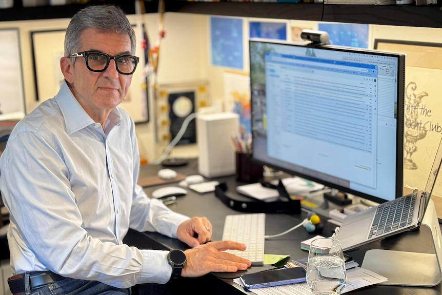 Daniel is older, Caucasian man, grey hair, looks seriously, sits in front of the computer, wears thick black-rimmed glasses.