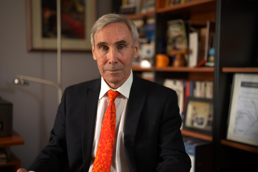 An older man with grey hair, wearing a suit and bright orange tie, looks neutrally into camera.